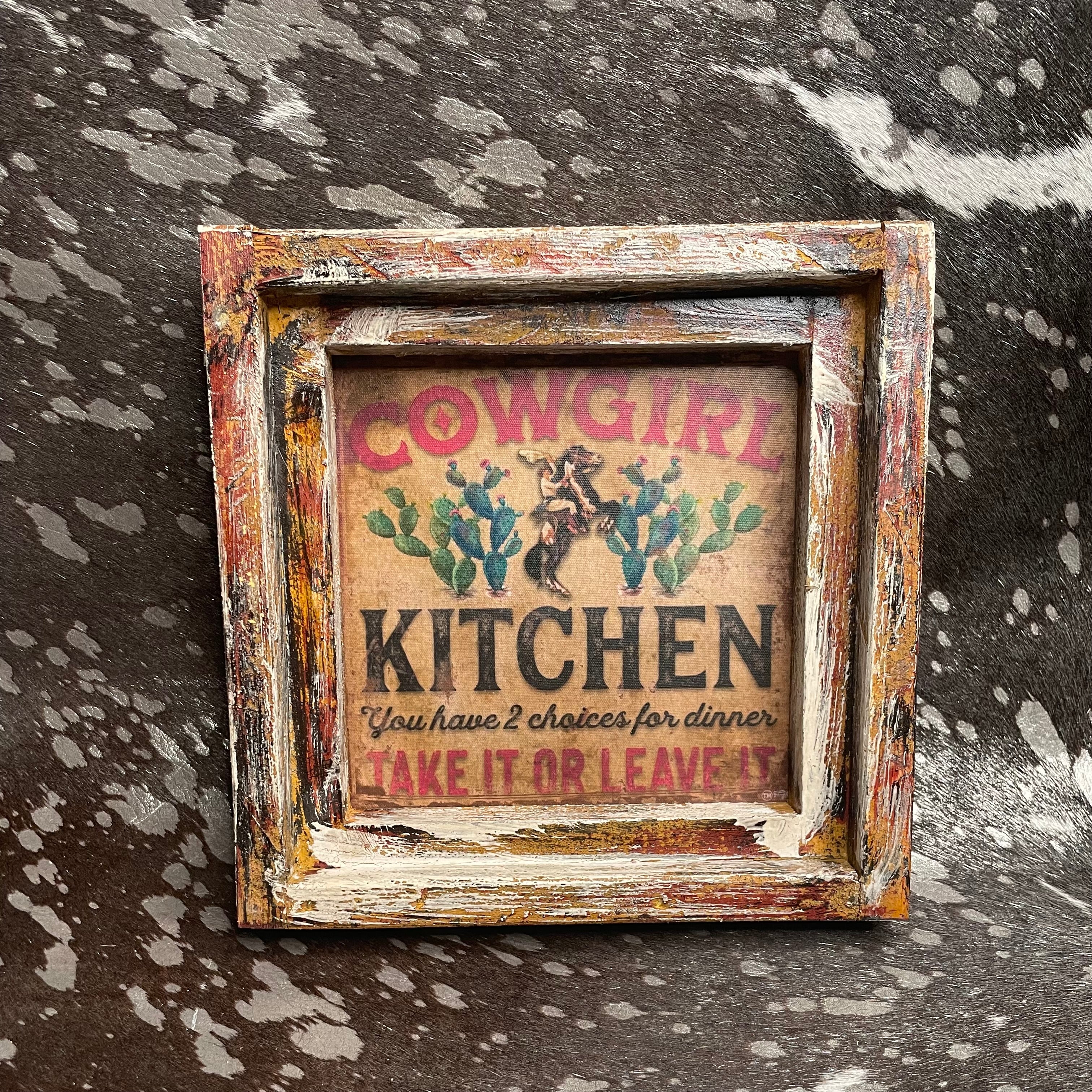 Decorative sign with 'Cowgirl Kitchen' text and graphics in a rustic frame on a textured surface.